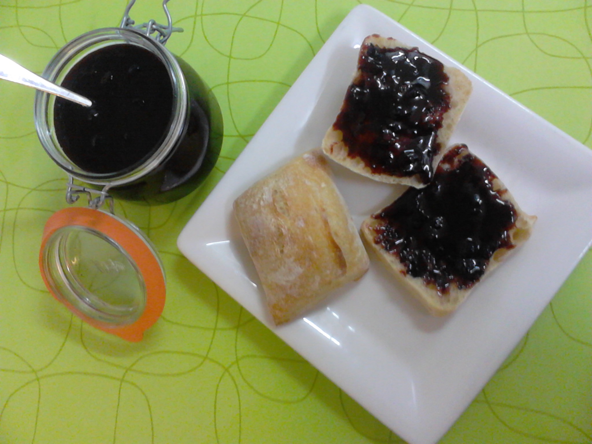 plate with two slices of bread spread with blackcurrant and wine jam, and a glass of blackcurrant and wine jam beside the plate