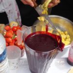 jug of exotic blackcurrant smoothie, someone cutting mangoes and strawberries in the background