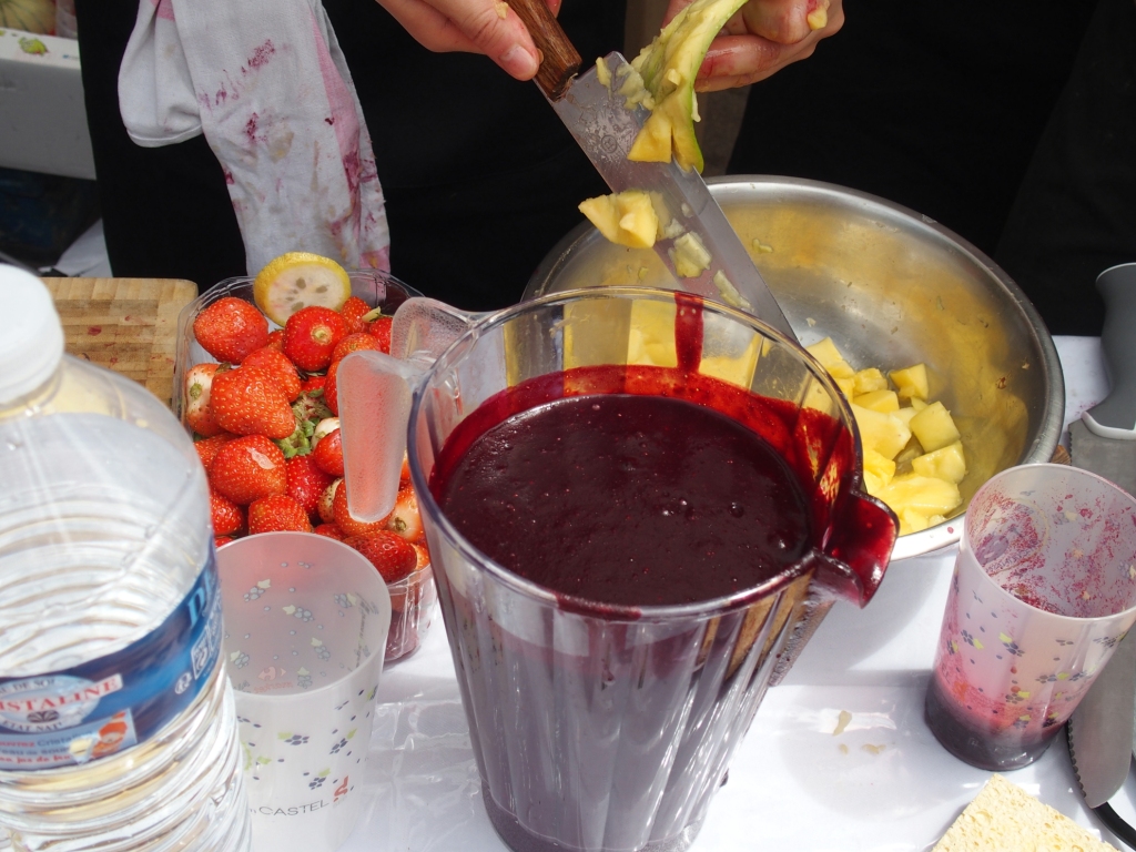 jug of exotic blackcurrant smoothie, someone cutting mangoes and strawberries in the background